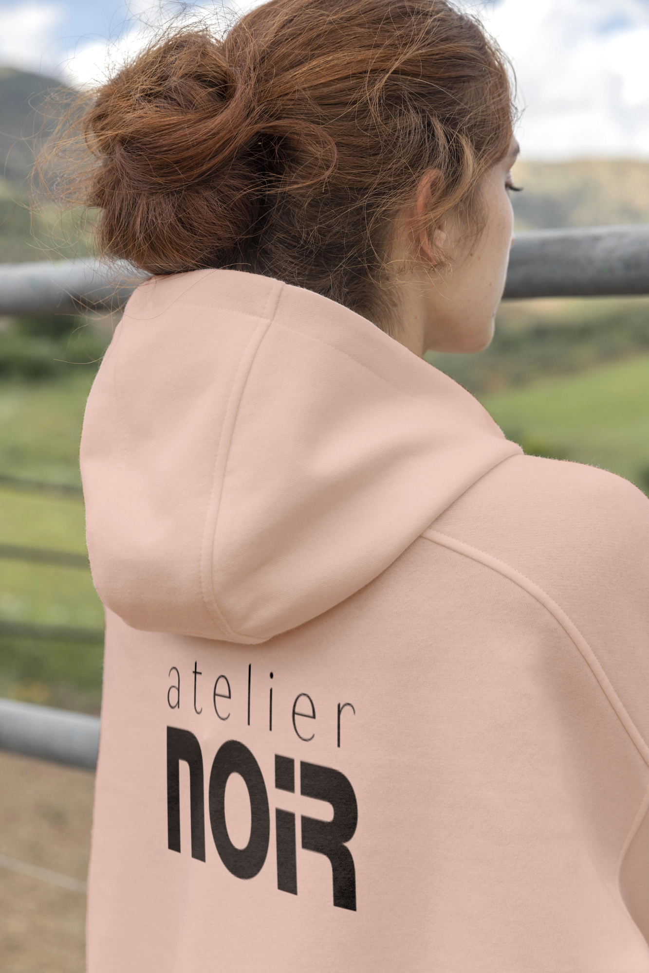 Вид сзади: женщина в бледно-розовой толстовке с логотипом Atelier NOIR.